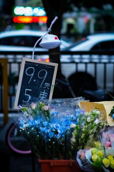 A display of fresh flower bouquets outdoors, with prominent blue and pink flowers arranged in clear wrapping, placed on a table or stand. Behind the flowers, there is a small chalkboard sign with hand
