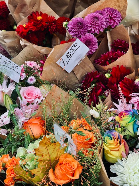 A vibrant assortment of freshly cut flowers including red dahlias, purple chrysanthemums, pink roses, and orange roses arranged in various brown paper bouquets. Some bouquets are wrapped with simple b
