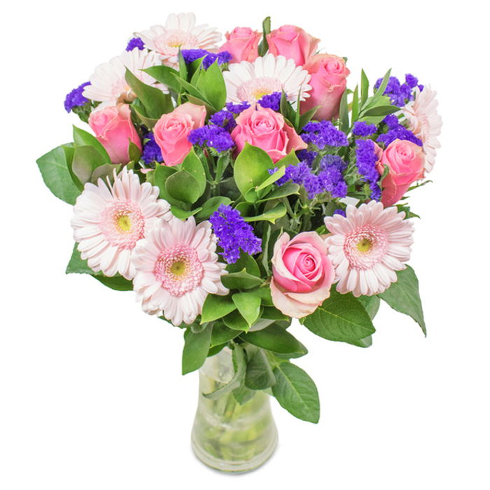 A vibrant, expertly arranged bouquet from New Addington Florist, gracefully presented in a clear glass vase that reveals fresh, carefully cut stems and sparkling water. This local arrangement captures the eye with a harmonious mix of 7 to 9 softly blushing pink roses, their petals either tightly furled or gently unfurling in a classic spiral, akin to the romance of a quiet evening stroll along King Henry's Drive. Around them, delicate light pink Gerbera daisies command attention with their broad, round faces and near-white centers, exuding a poised yet playful charm. Interwoven among the main blooms, bold clusters of deep purple statice infuse the design with a splash of vivid colour and intriguing texture, reminiscent of the lively hues seen at community flower shows. A generous base of rich, leafy greenery grounds the bouquet, offering a sense of abundance and reinforcing the freshness so valued by New Addington residents. The entire arrangement feels both joyful and composed, suitable for celebrations such as birthdays, anniversaries, or simply as a lovely accent for a well-lit sitting room overlooking Addington Park. Conjuring feelings of affection and joy, this bouquet brings a gentle celebration of natural beauty-and the care of skilled local hands-right into your home.